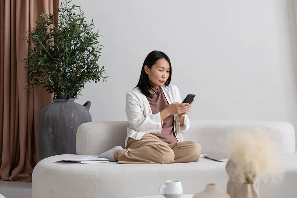 A woman sitting cross-legged on a pale sofa, looking at her phone, with a notebook and laptop beside her