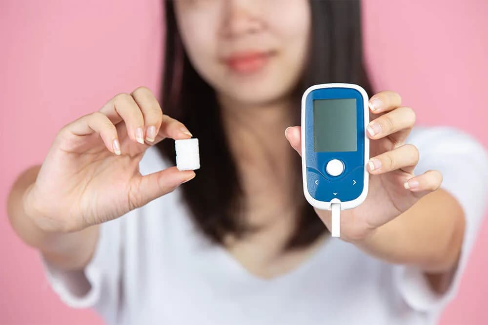 A woman holding a sugar cube in one hand and a blood glucose meter in the other, both toward the camera