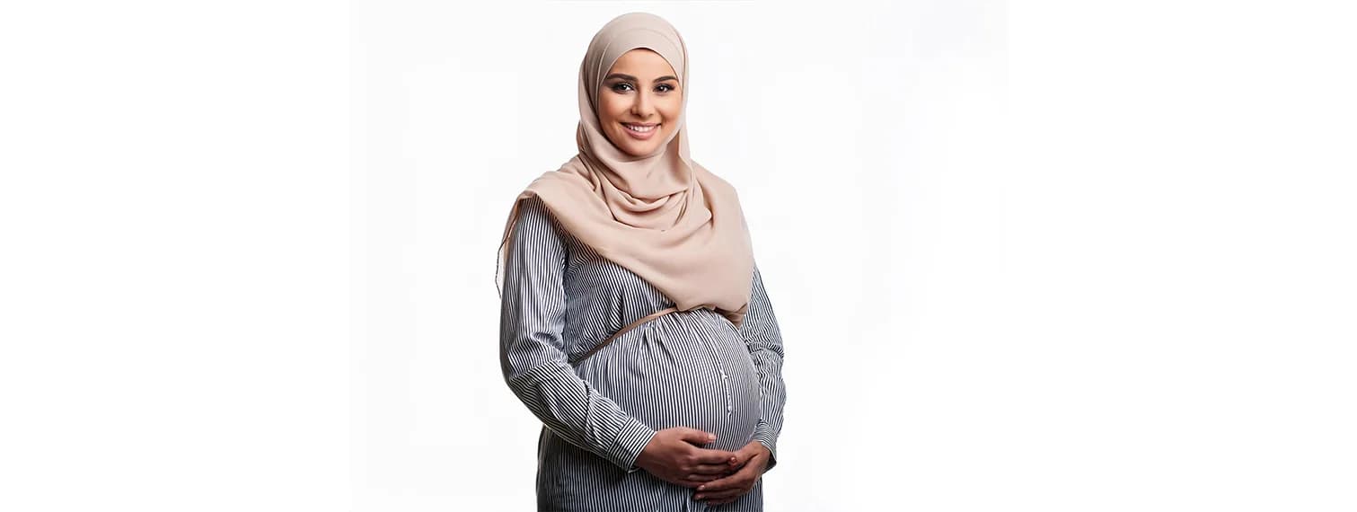 امرأة حامل مبتسمة بحجاب بيج وقميص مخطط تحتضن بطنها على خلفية بيضاء