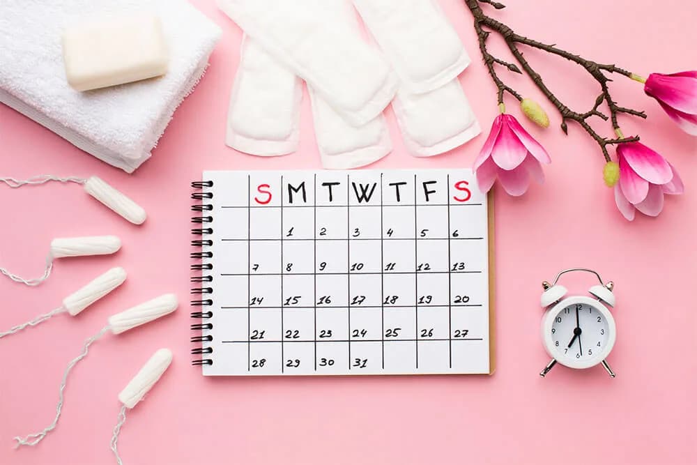 Menstrual pads and tampons, a bar of soap and a small alarm clock arranged around a spiral notepad calendar on a pink background