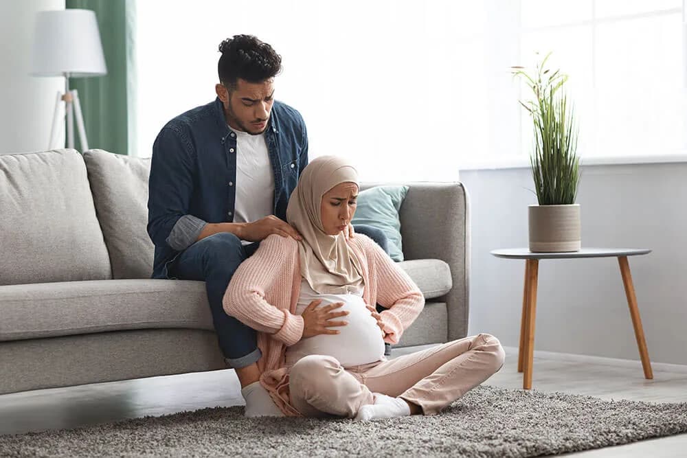 A pregnant woman in a hijab sitting on the floor holding her abdomen with a pained expression while a man beside her rests a hand on her shoulder