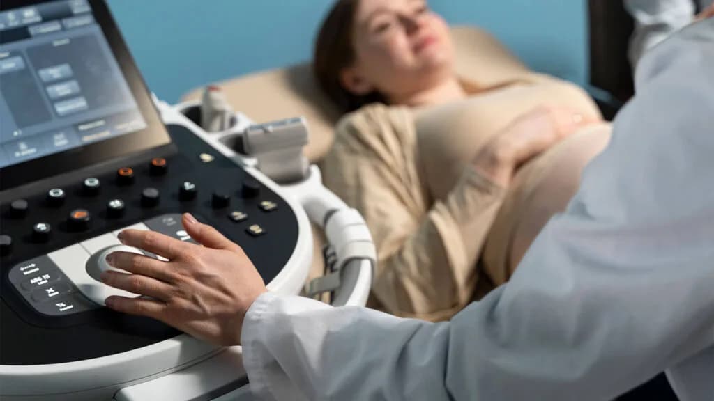 A clinician operating an ultrasound console while a woman lies on the examination couch behind it