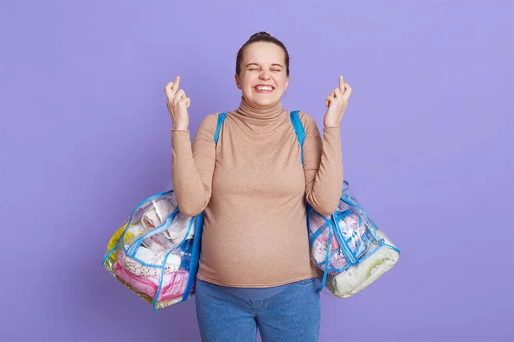 A pregnant woman against a lilac background laughing with her fingers crossed, a packed transparent hospital bag hanging from each shoulder
